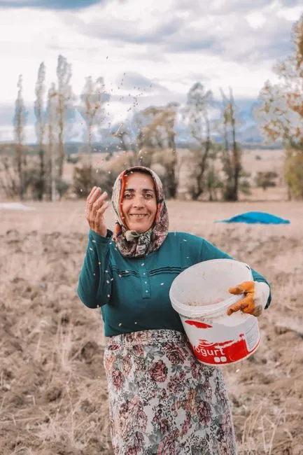 Une femme joyeuse dans un cadre rural répand des graines dans le champ, transmettant tradition et agriculture.