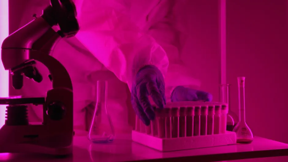 Scientifique en blouse de laboratoire manipulant des tubes à essai sous un éclairage rose, utilisant un microscope.